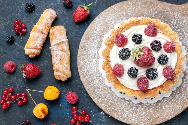 Draufsicht auf köstlichen kleinen Kuchen mit Zuckerpulvercreme und Beeren zusammen mit Armreifen auf dunklem Schreibtisch