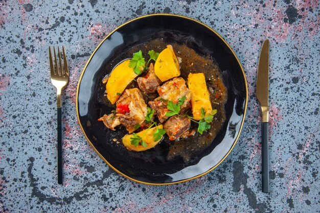 Draufsicht auf köstliches Abendessen mit Fleischkartoffeln, die mit Grün in einem schwarzen Teller und Besteck auf Mischfarbenhintergrund gesetzt werden