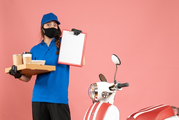 Draufsicht auf Kuriermädchen mit medizinischen Maskenhandschuhen, die neben dem Motorrad stehen und Dokumente und Kaffeekuchen auf pastellfarbenem Hintergrund in Pfirsichfarbe halten