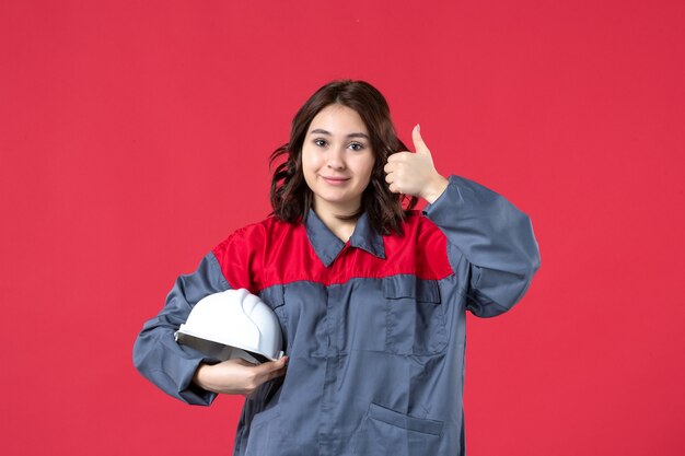 Draufsicht auf lächelnde Baumeisterin in Uniform und mit Schutzhelm, die eine ok Geste auf isoliertem rotem Hintergrund macht