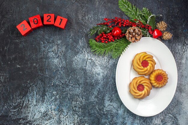 Draufsicht auf leckere Kekse auf einem weißen Teller und Neujahrsdekorationen Weihnachtsmann-Hut neben Zahlen auf dunkler Oberfläche