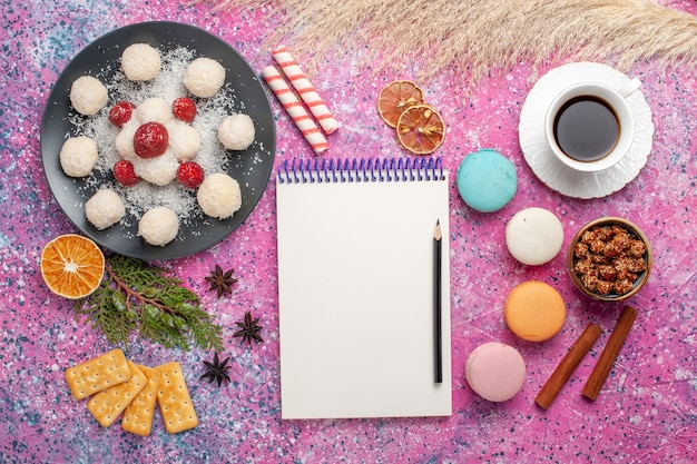 Draufsicht auf leckere Kokosnussbonbons mit Macarons und Tasse Tee auf hellrosa Oberfläche