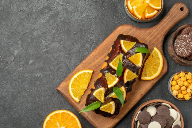 Draufsicht auf leckere Kuchen ganz und geschnittene Orangen mit Keksen auf Schneidebrett auf dunklem Tisch