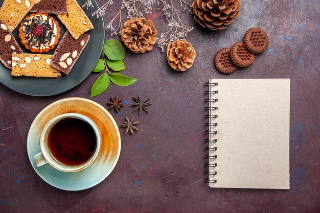 Draufsicht auf leckere Kuchenscheiben mit einer Tasse Tee und Keksen auf Schwarz