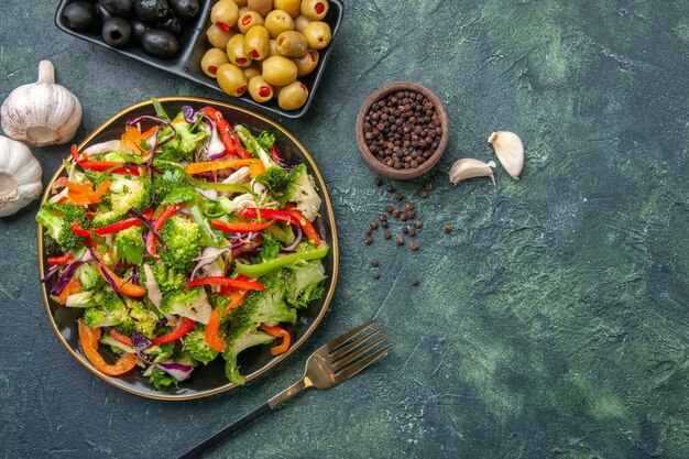 Draufsicht auf leckeren veganen Salat in einem Teller mit verschiedenem Gemüse und Gabelpfeffer grüne schwarze Oliven Knoblauch auf der rechten Seite auf dunklem Hintergrund