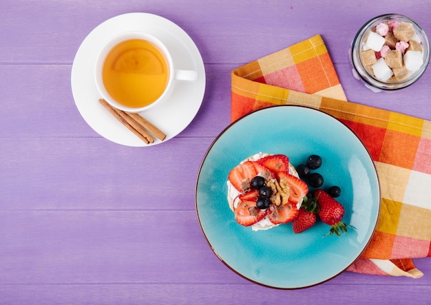 Draufsicht auf leckeres Knäckebrot mit reifen Blaubeeren, Erdbeeren und Nüssen mit saurer Sahne auf einer Keramikplatte, serviert mit einer Tasse grünem Tee auf lila Holzhintergrund