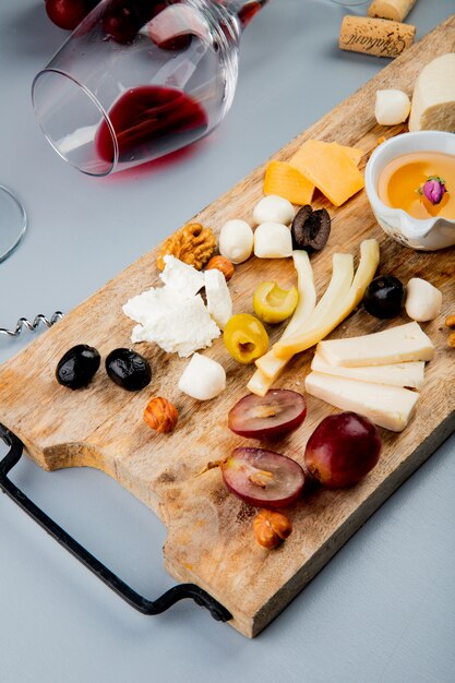 Draufsicht auf liegendes Glas Rotwein mit verschiedenen Arten von Käsetrauben-Oliven-Nussbutter auf Schneidebrett und Kork auf Weiß 1