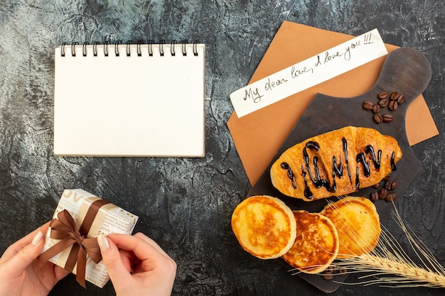 Kostenloses Foto draufsicht auf notizbuch und leckeres frühstück mit pfannkuchen-croisasant und handöffnender geschenkbox auf dunklem tisch