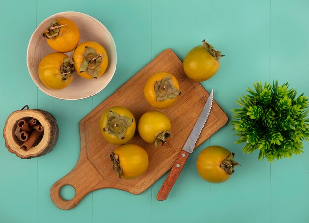 Draufsicht auf orangefarbene rundliche Kakifrüchte auf einem hölzernen Küchenbrett mit Messerzimtstangen auf einem Holzglas auf einem blauen Holztisch