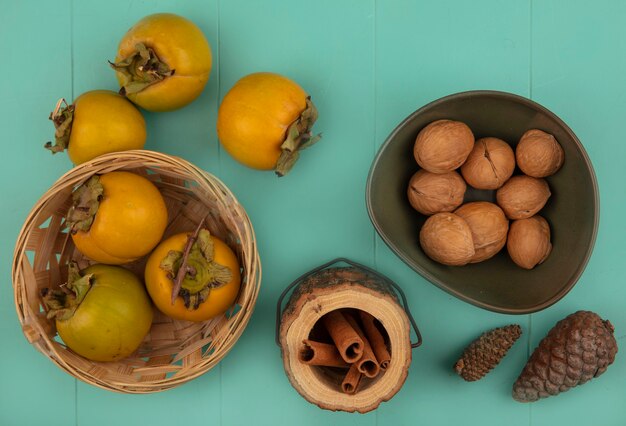 Draufsicht auf orangefarbene rundliche Persimonenfrüchte auf einem Eimer mit Zimtstangen auf einem Holzglas mit Walnüssen auf einer Schüssel mit Persimonenfrüchten, die auf einem blauen Holztisch lokalisiert werden