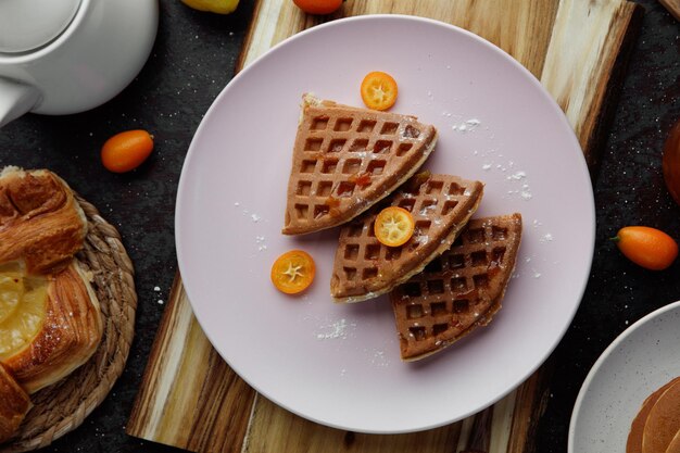 Draufsicht auf Pfannkuchen mit Kumquat-Scheiben in Teller auf Schneidebrett mit Croissant, ganzen Kumquats und Teekanne auf schwarzem Hintergrund