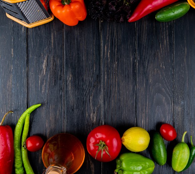 Draufsicht auf Reibe und Flasche Olivenöl mit frischem Gemüse grüne Chilischoten Tomaten Gurken bunte Paprika und Zitrone auf dunklem Holz mit Kopienraum