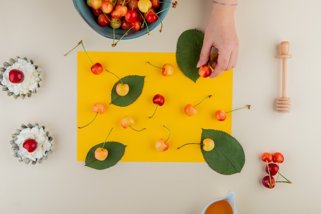 Draufsicht auf reife regnerischere Kirschen auf einem Blatt gelben Papiers und Hüttenkäse in Mini-Tortenformen auf Weiß