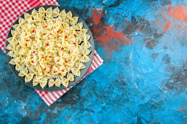 Draufsicht auf rohe italienische Farfalle-Nudeln mit Gemüse auf einem schwarzen Teller auf rot gestreiftem Handtuch auf der rechten Seite auf blauem Hintergrund