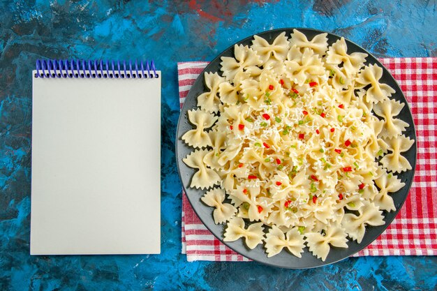 Draufsicht auf rohe italienische Farfalle-Nudeln mit Gemüse auf rotem, abgestreiftem Handtuch neben geschlossenem Notizbuch auf blauem Tisch