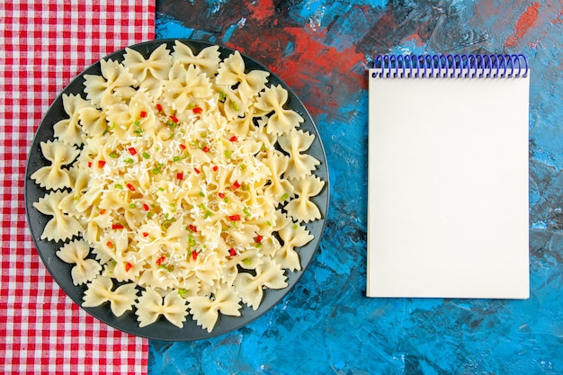 Draufsicht auf rohe italienische Farfalle-Nudeln mit Gemüse auf rotem, abgestreiftem Handtuch und Spiralnotizbuch auf blauem Tisch