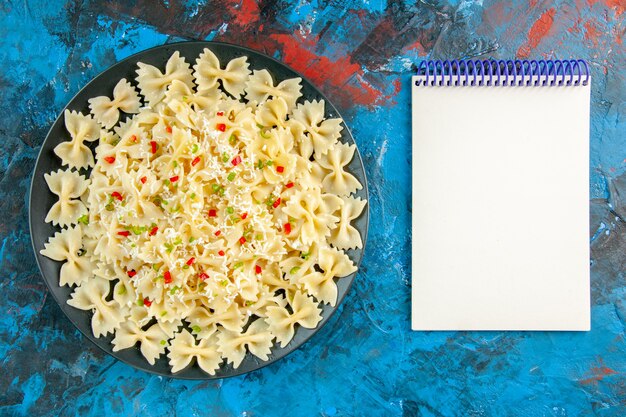 Draufsicht auf rohe italienische Farfalle-Nudeln mit Gemüse und Spiralnotizbuch auf blauem Hintergrund