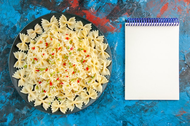 Draufsicht auf rohe italienische Farfalle-Nudeln mit Gemüse und Spiralnotizbuch auf blauem Hintergrund