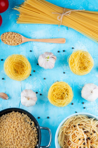 Draufsicht auf rohe Nudeln verschiedener Formen und Arten als Spaghetti-gelbes Nudelnest und sternförmige Nudeln mit frischen Tomaten und Knoblauch auf Blau