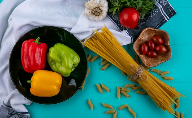 Draufsicht auf rohe Spaghetti mit glockenfarbenen Paprika-Tomaten und Knoblauch auf einer türkisfarbenen Oberfläche