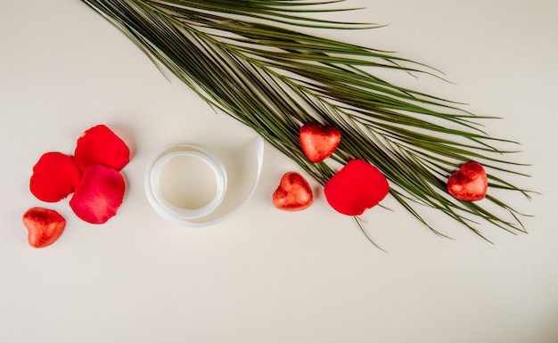 Draufsicht auf rote Rosenblätter, herzförmige Pralinen, eingewickelt in rote Folie und Palmblatt mit Band auf weißem Tisch