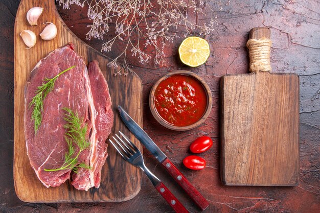 Draufsicht auf rotes Fleisch auf Holzbrett und Ketchup in kleiner Schüsselgabel und Messer auf dunklem Hintergrund stockbild