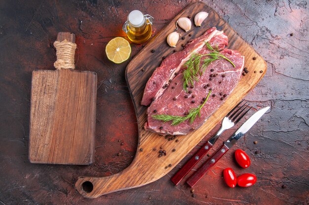 Draufsicht auf rotes Fleisch auf Holzbrett und Knoblauch grüner Pfeffer oi Flaschengabel und Messer auf dunklem Hintergrund