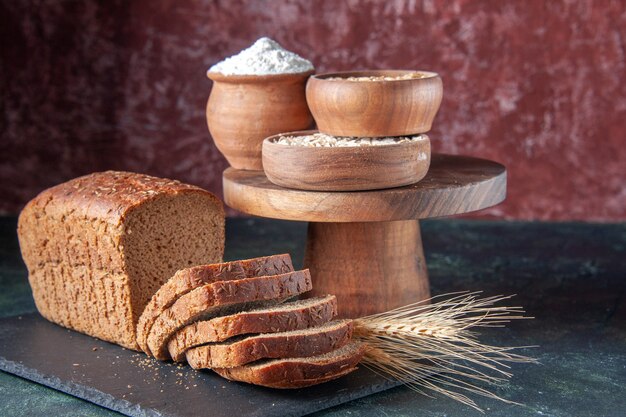 Draufsicht auf Schwarzbrotscheiben auf dunklem Farbtablett Mehl Haferflocken Buchweizen auf Holzbrett auf gemischten Farben beunruhigtem Hintergrund