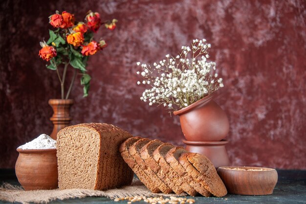 Draufsicht auf Schwarzbrotscheiben Mehl in einer Schüssel und Weizen auf nacktem Farbtuch und Blumentöpfen auf Mischfarbenhintergrund