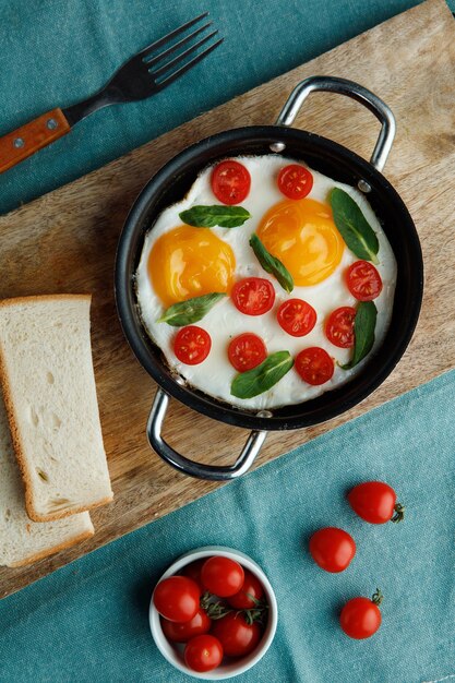 Draufsicht auf Spiegeleier mit Tomaten, dekoriert mit Blättern in der Pfanne mit Brotscheiben auf Schneidebrett und Tomaten mit Gabel auf blauem Hintergrund