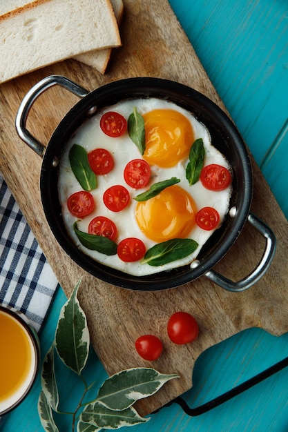 Draufsicht auf Spiegeleier mit Tomaten in Bratpfanne, dekoriert mit Blättern mit Brotscheiben auf Schneidebrett mit Tasse Orangensaft-Grapefruit auf kariertem Stoff auf blauem Hintergrund