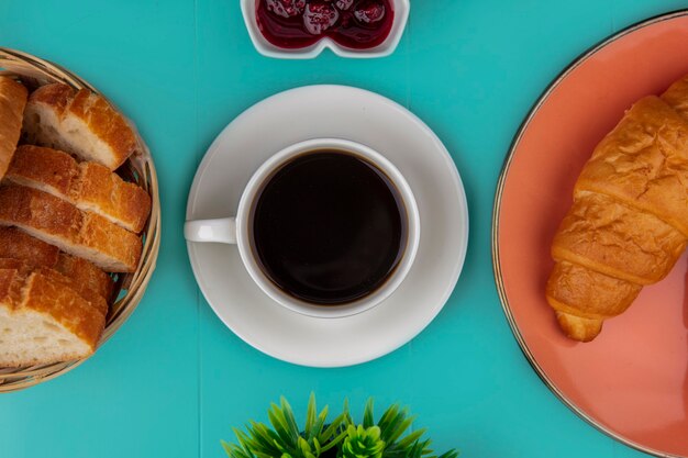 Draufsicht auf Tasse Tee und Croissant geschnittenes Baguette mit Himbeermarmelade und Pflanze auf blauem Hintergrund