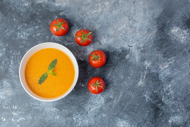 Draufsicht auf Tomatensuppe in weißer Keramikschale mit frischen Tomaten auf grauem Tisch.