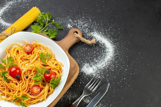 Draufsicht auf vegane spagetti-mahlzeit mit tomaten und grün auf einem quadratischen teller rohe nudeln, die von einem seilbesteck auf der rechten seite auf schwarzweißem hintergrund gefesselt sind