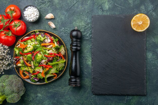 Draufsicht auf veganen Salat in einem Teller mit verschiedenem Gemüse und Gabeltomaten mit schwarzem Hammerknoblauch-Brokolli-Blumenschneidebrett auf dunklem Hintergrund