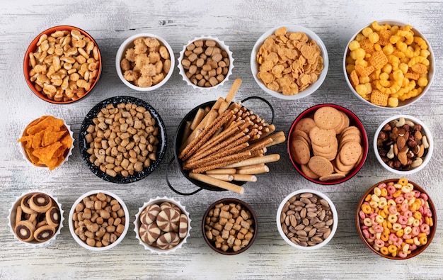 Draufsicht auf verschiedene Arten von Snacks als Nüsse, Cracker und Kekse in Schalen auf weißer Holzoberfläche horizontal