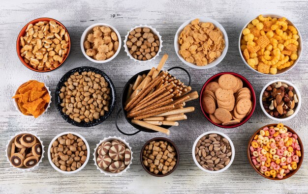 Draufsicht auf verschiedene Arten von Snacks als Nüsse, Cracker und Kekse in Schalen auf weißer Holzoberfläche horizontal