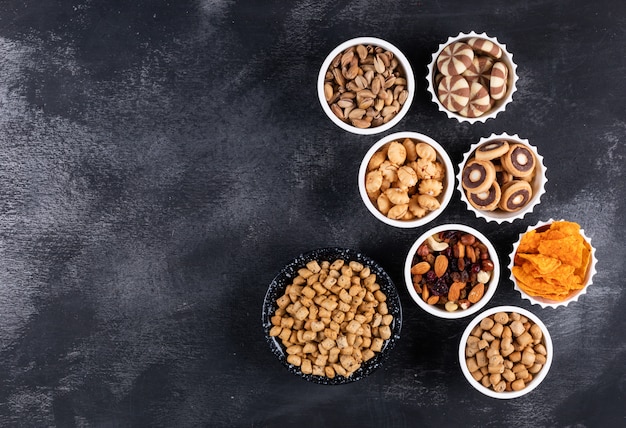 Draufsicht auf verschiedene Arten von Snacks als Nüsse, Cracker und Kekse in Schalen mit Kopienraum auf dunklem Hintergrund horizontal