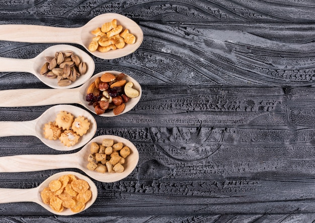 Draufsicht auf verschiedene Arten von Snacks als Nüsse und Cracker auf Holzlöffeln mit Kopienraum auf dunklem Hintergrund horizontal