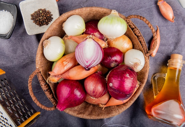 Draufsicht auf verschiedene Arten von Zwiebeln im Korb mit Salzschwarzpfeffersamen geschmolzene Butterreibe herum auf grauem Stoff