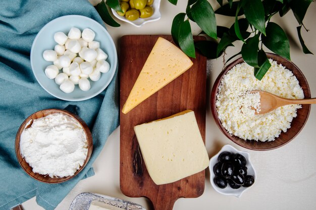 Draufsicht auf verschiedene Käsesorten auf Holzschneidebrett und Hüttenkäse in einer Holzschale mit eingelegten Oliven auf rustikalem Tisch