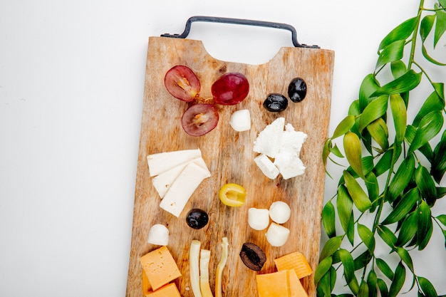 Kostenloses Foto draufsicht auf verschiedene käsesorten mit traubenstücken und oliven auf schneidebrett auf weiß, verziert mit blättern mit kopierraum