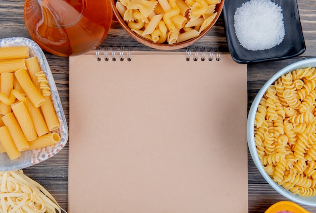 Draufsicht auf verschiedene Makkaronis als ziti rotini tagliatelle und andere mit geschmolzenem Buttersalz um Notizblock auf Holz mit Kopierraum