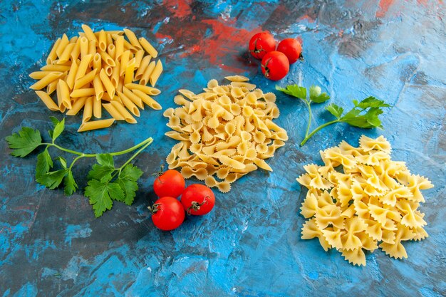 Draufsicht auf verschiedene rohe italienische Pasta in einer Reihe und Tomaten mit Stiel auf blauem Hintergrund