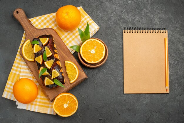 Draufsicht auf weiche Kuchen ganz und geschnittene Zitronen mit Blättern neben Notizbuch auf dunklem Tisch