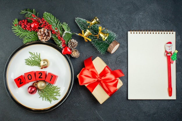 Draufsicht auf Zahlendekorationszubehör auf einer Platte Tannenzweige Nadelbaumkegel-Weihnachtsbaum-Notizbuch mit Stift auf dunklem Hintergrund