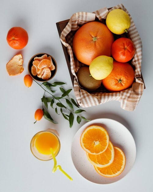 Draufsicht auf Zitrusfrüchte als Orangen-Mandarinen-Zitronen-Kiwi in Schachtel mit Mandarinen- und Orangenscheiben in Schüssel und Teller mit Orangensaft und Mandarinenschale mit Blättern auf weißem Hintergrund