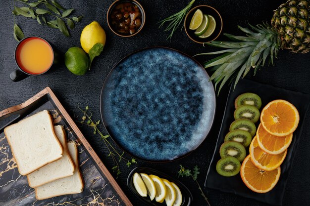 Draufsicht auf Zitrusfrüchte wie Ananas-Kiwi-Orange-Limetten-Zitronen- und Mandarinenscheiben mit Brotscheiben in Tablett und Marmelade in Schüssel um leeren Teller auf schwarzem Hintergrund