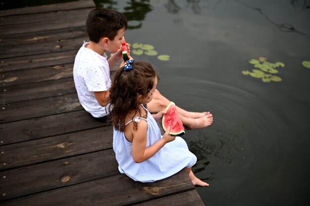 Draufsicht auf zwei kinder, die auf dem pier sitzen, ihre füße mit seerosen im teich senken, sich in der natur entspannen, an der frischen luft, frische reife wassermelone essen
