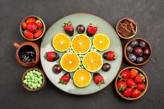 Draufsicht aus der Ferne Beeren und Schokoladenteller mit schokoladenüberzogenen Erdbeeren und gehackter Orange neben den Schalen mit Schokoladensauce Süßigkeiten und Beeren auf dem Tisch